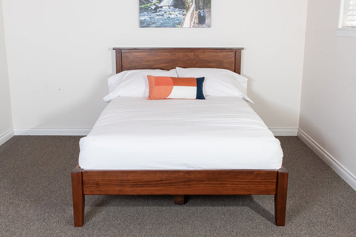 A solid wood platform bed frame with a headboard, displayed in a room with a white mattress and bedding, in a brown stain finish.