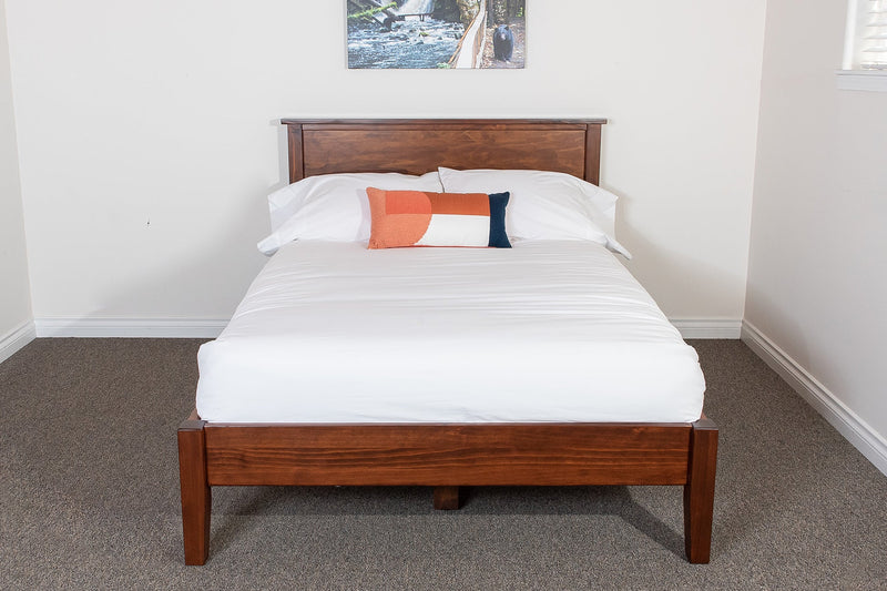 A solid wood platform bed frame with a headboard, displayed in a room with a white mattress and bedding, in a brown stain finish.