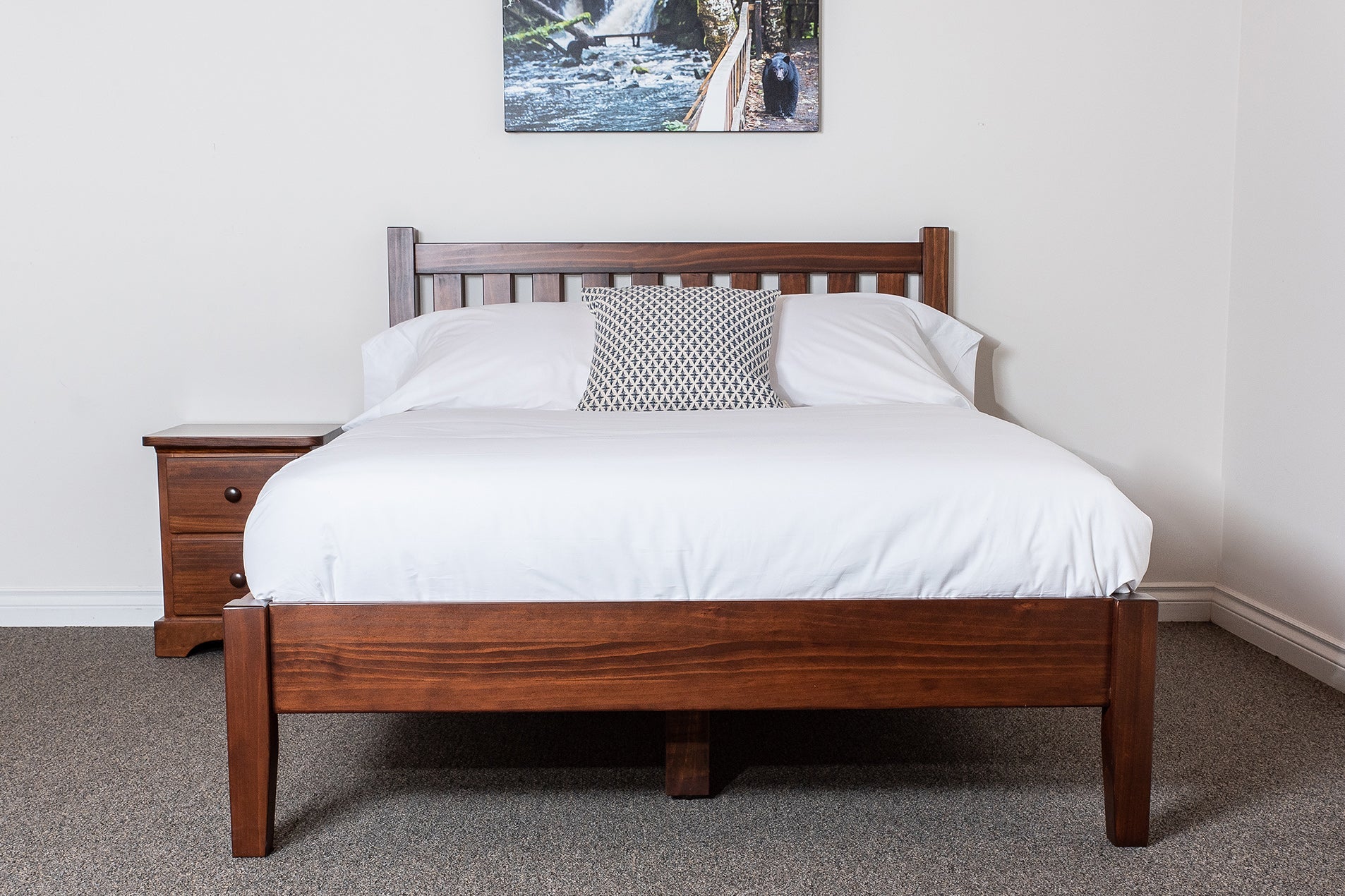 A slatted platform bed frame made of pine with a white mattress, brown slats, and a wooden headboard. The bed is displayed in a room setting with a nightstand on the left and a framed picture above the bed.